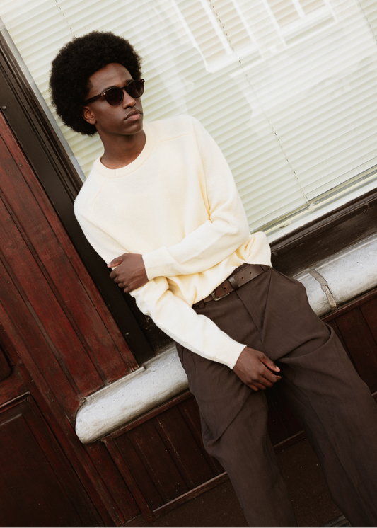A man with an afro hairstyle wearing dark sunglasses, a cream-colored long-sleeve crewneck sweater, and brown trousers, leaning against a dark wooden door next to a window with white blinds.